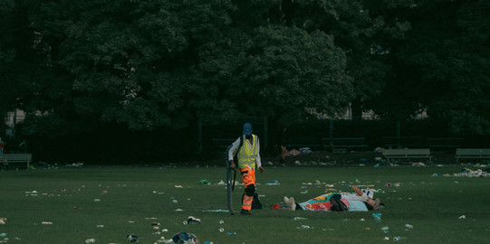 worker picking up garbage on a field next to two people lying down