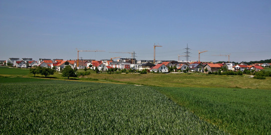 Baugebiet am Stadtrand mit Ackerfläche im Vordergrund und Baufläche für Gebäude mit Baukränen im Hintergrund