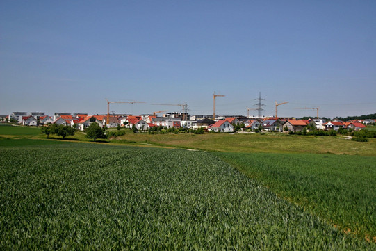 Baugebiet am Stadtrand mit Ackerfläche im Vordergrund und Baufläche für Gebäude mit Baukränen im Hintergrund